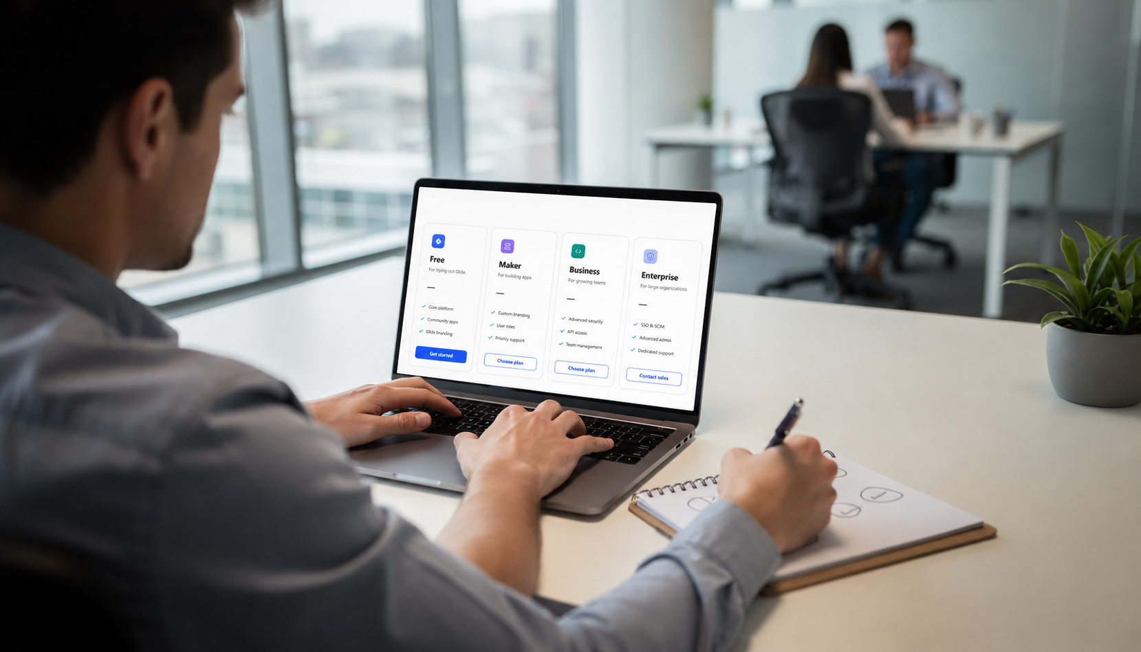 A person is seated at a modern office desk, intently comparing various glide pricing plans for software on their laptop screen, which displays options like business plans and free trials. The environment features a sleek design, emphasizing productivity and the use of no code tools for building apps.